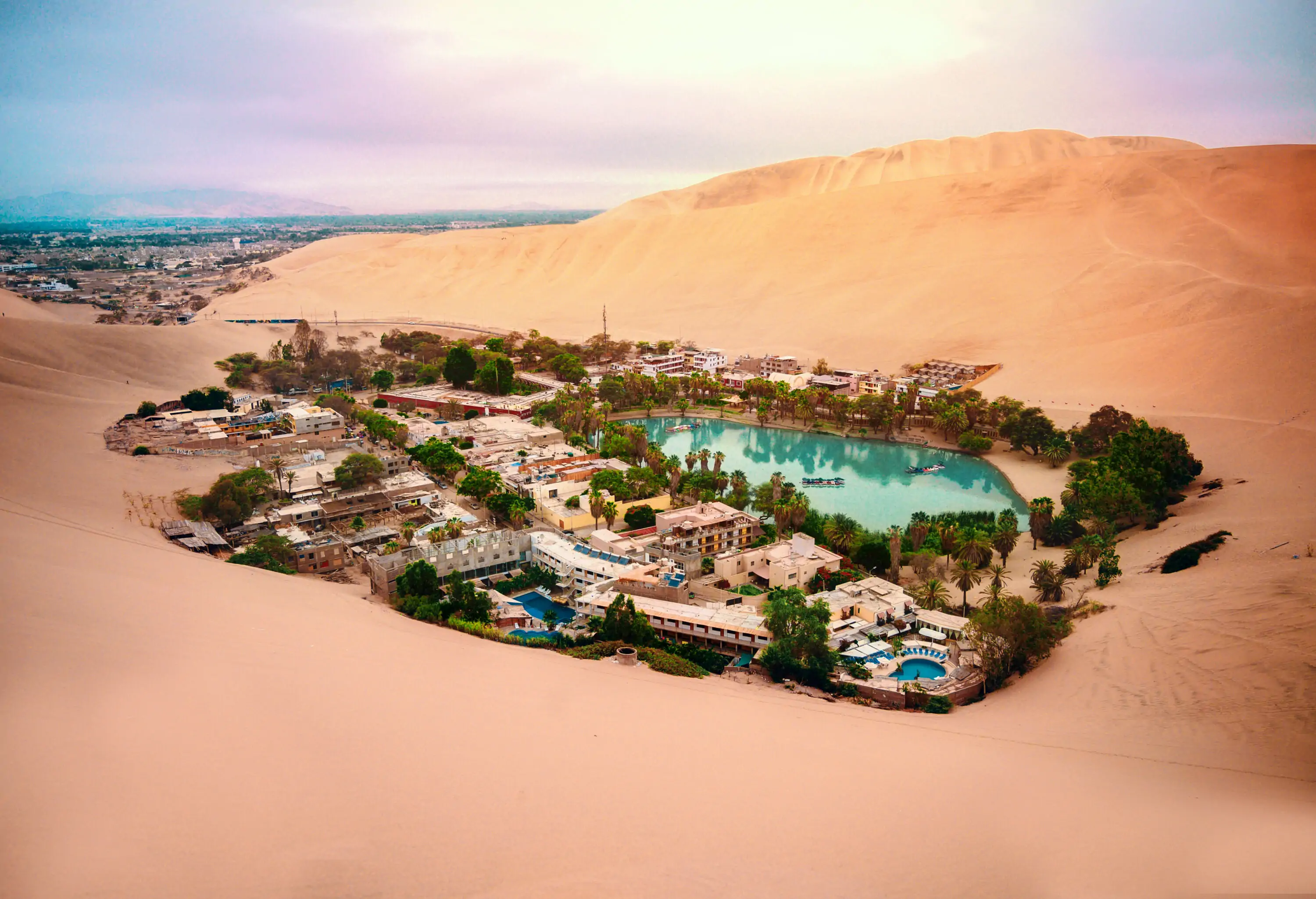 A cluster of buildings amongst lush trees beside a small pond in an oasis surrounded by sand dunes.