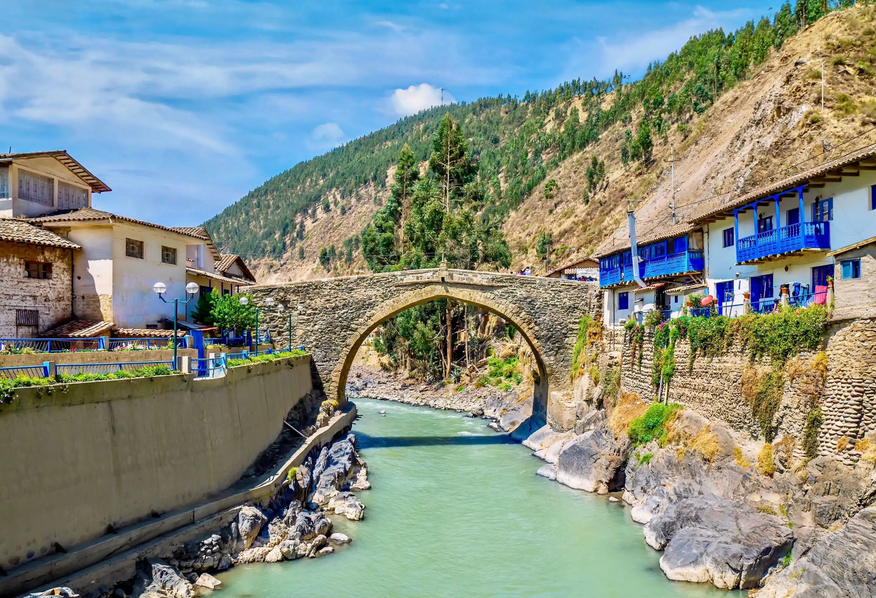 An old, historic, European-style arched bridge made of stone, spanning a river running through a quaint, rustic Peruvian town, with buildings on both sides, some with decorative blue balconies.